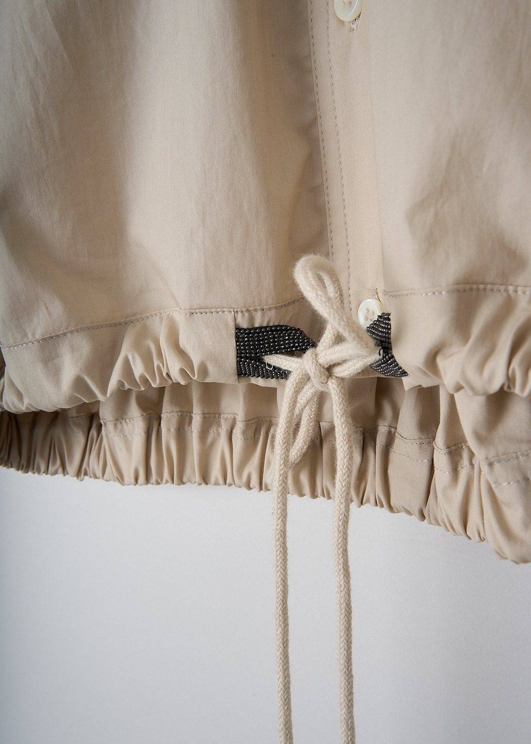 Brunello Cucinelli, Cropped beige blouse with drawstring hem, M0H93NE616_C8617, Beige, Detail, This beige short-sleeved blouse has a classic shirt collar and a front button closure. The short sleeves have straight hems. In the front, the blouse has two breast pockets. The blouse has partly elasticated hem with a pleated drawstring and subtle monili detailing. In the back, the blouse has a single vertical box pleat. The blouse has a cropped length. 

