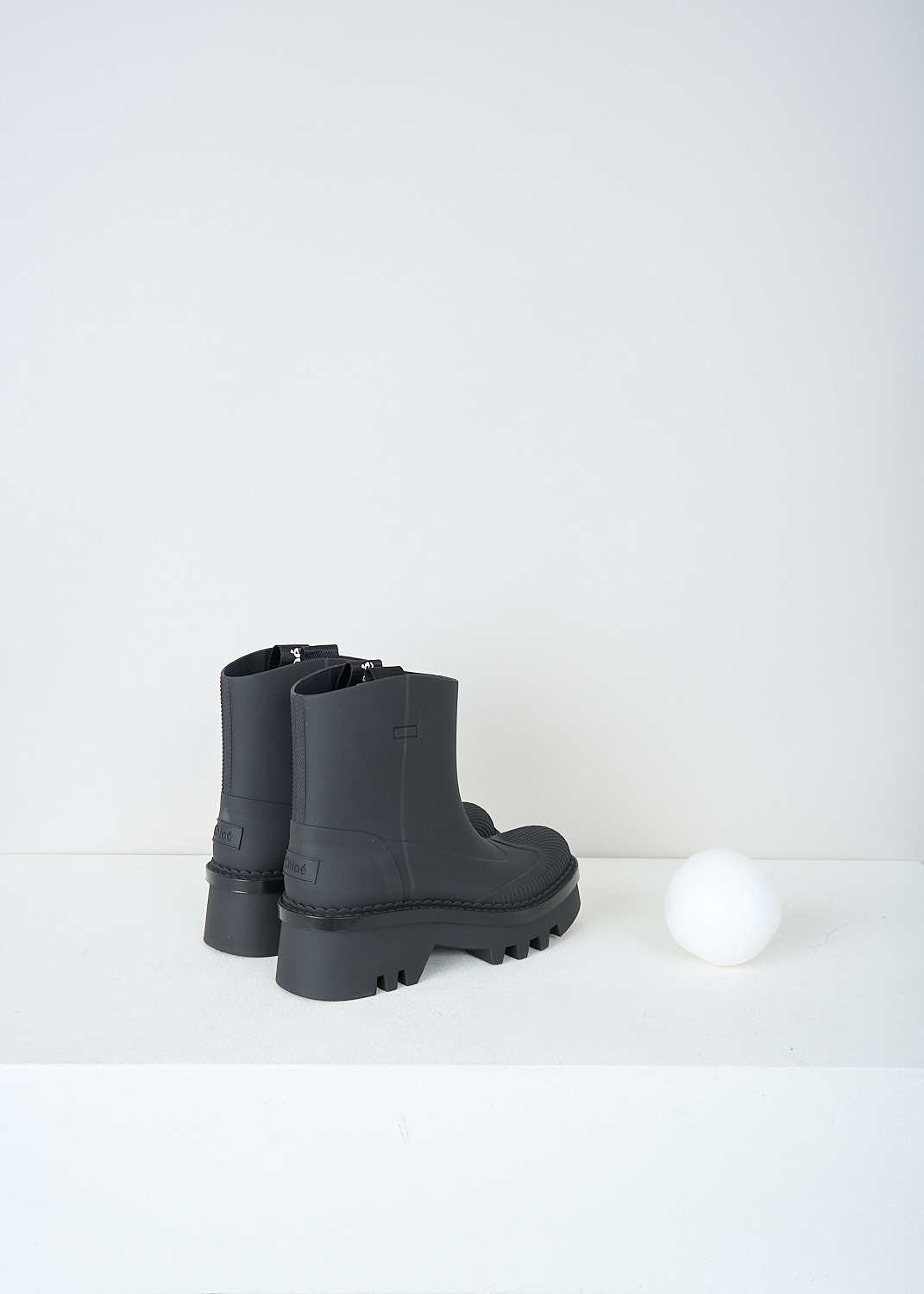 Chloé, Chunky short black rain boots, CHC23A904FP001_RAINBOOTS_BLACK, Black, Back, These chunky short black slip-on rain boots have two pull tabs on the top edge with the brand's name on them in white lettering. These boots have a round toe with ribbed detailing. A subtle scalloped trim runs along the chunky ridged sole.  
