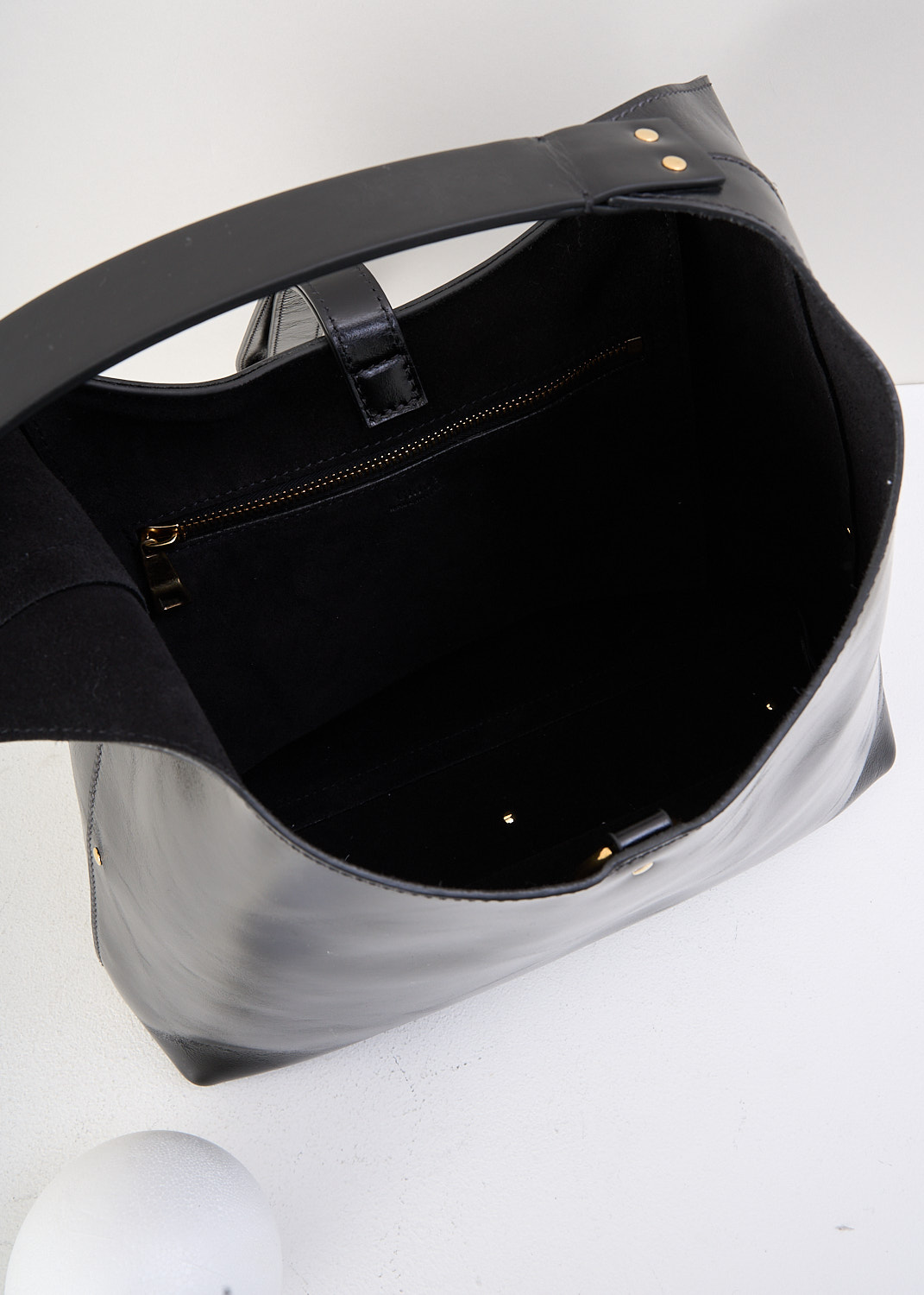 Chloé, Marcie tote bag in black, CHC24AS629N57_001_BLACK, Black, Detail 1, This Marcie tote bag is made from supple black leather with a smooth, subtly glossy finish. It features a soft, slouchy, crescent-shaped silhouette and a single shoulder strap. Gold-tone hardware is used throughout. The bag closes with a vertical leather strap with small metal-tipped ends and a gold-tone O-ring at the top. Inside, the bag has a zipped slip pocket. 
