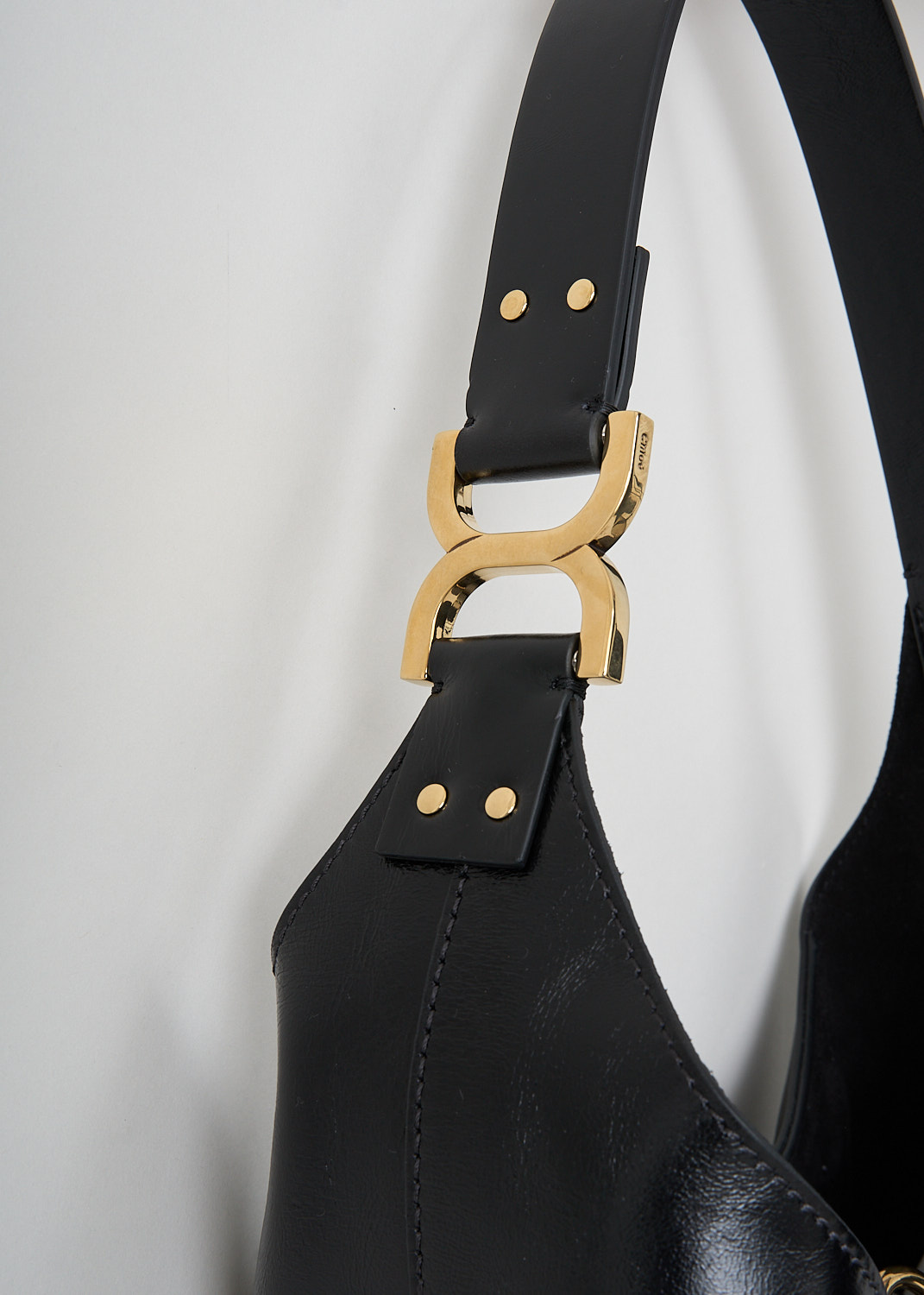 Chloé, Marcie tote bag in black, CHC24AS629N57_001_BLACK, Black, Detail, This Marcie tote bag is made from supple black leather with a smooth, subtly glossy finish. It features a soft, slouchy, crescent-shaped silhouette and a single shoulder strap. Gold-tone hardware is used throughout. The bag closes with a vertical leather strap with small metal-tipped ends and a gold-tone O-ring at the top. Inside, the bag has a zipped slip pocket. 
