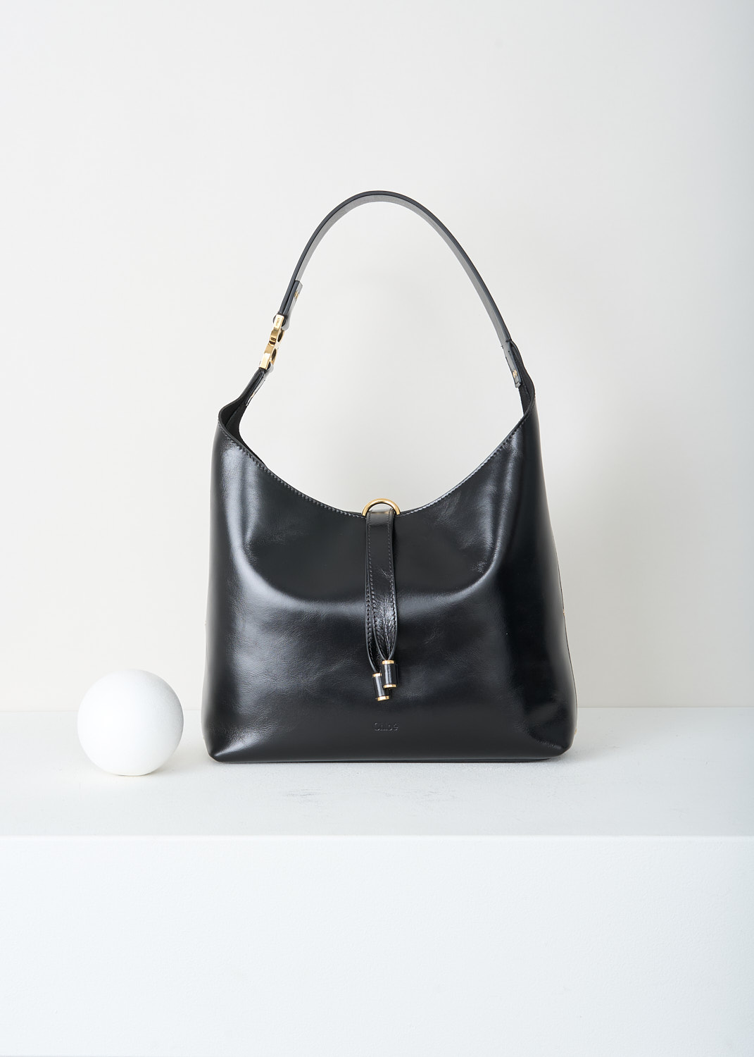 Chloé, Marcie tote bag in black, CHC24AS629N57_001_BLACK, Black, Front, This Marcie tote bag is made from supple black leather with a smooth, subtly glossy finish. It features a soft, slouchy, crescent-shaped silhouette and a single shoulder strap. Gold-tone hardware is used throughout. The bag closes with a vertical leather strap with small metal-tipped ends and a gold-tone O-ring at the top. Inside, the bag has a zipped slip pocket. 
