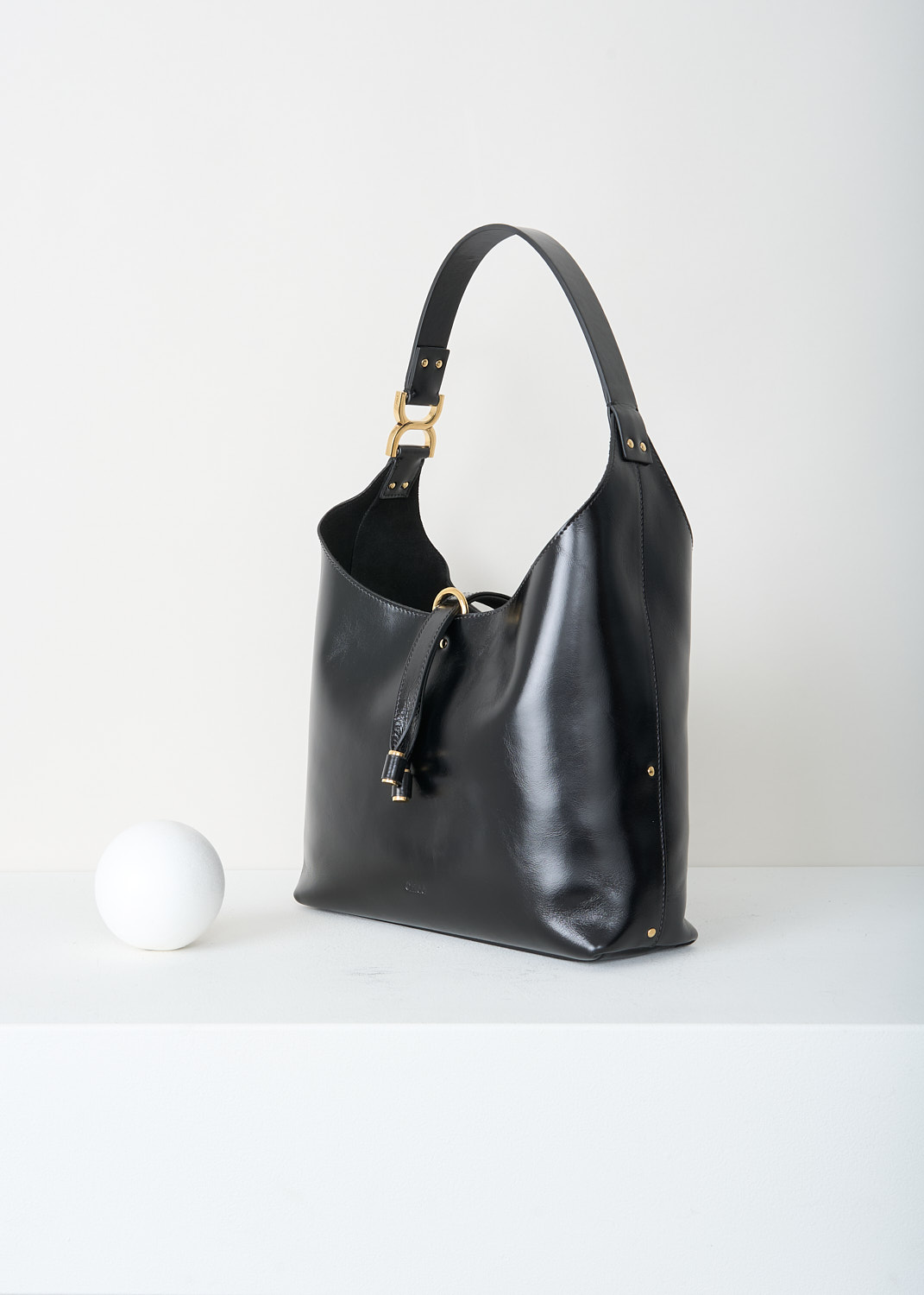 Chloé, Marcie tote bag in black, CHC24AS629N57_001_BLACK, Black, Side, This Marcie tote bag is made from supple black leather with a smooth, subtly glossy finish. It features a soft, slouchy, crescent-shaped silhouette and a single shoulder strap. Gold-tone hardware is used throughout. The bag closes with a vertical leather strap with small metal-tipped ends and a gold-tone O-ring at the top. Inside, the bag has a zipped slip pocket. 
