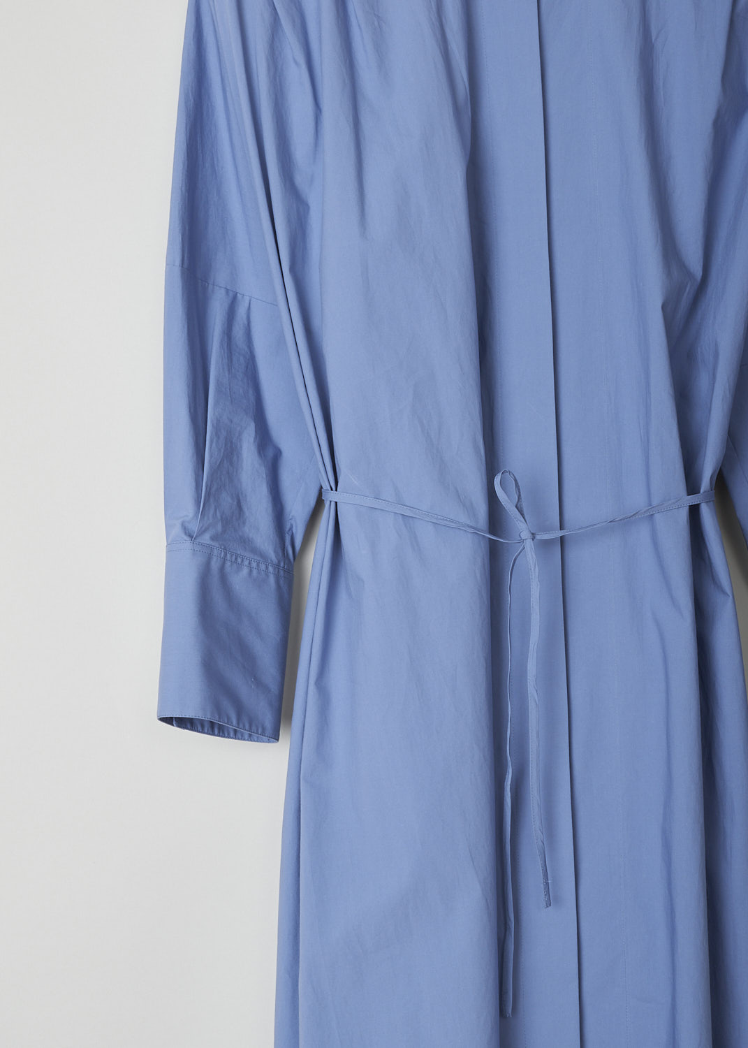 JIL SANDER, BLUE MIDI SHIRT DRESS WITH WAIST TIE, JSPO500806_WO242000_450, Blue, Detail, This blue midi shirt dress has a spread collar with a straight neckline. The dress has a concealed front button closure with a single black button visible. The dress has three quarter sleeves with buttoned cuffs. The A-line silhouette can be cinched in with ties. Slanted pockets are concealed in the side seams. The dress has an asymmetrical finish, meaning the back is longer than the front.
