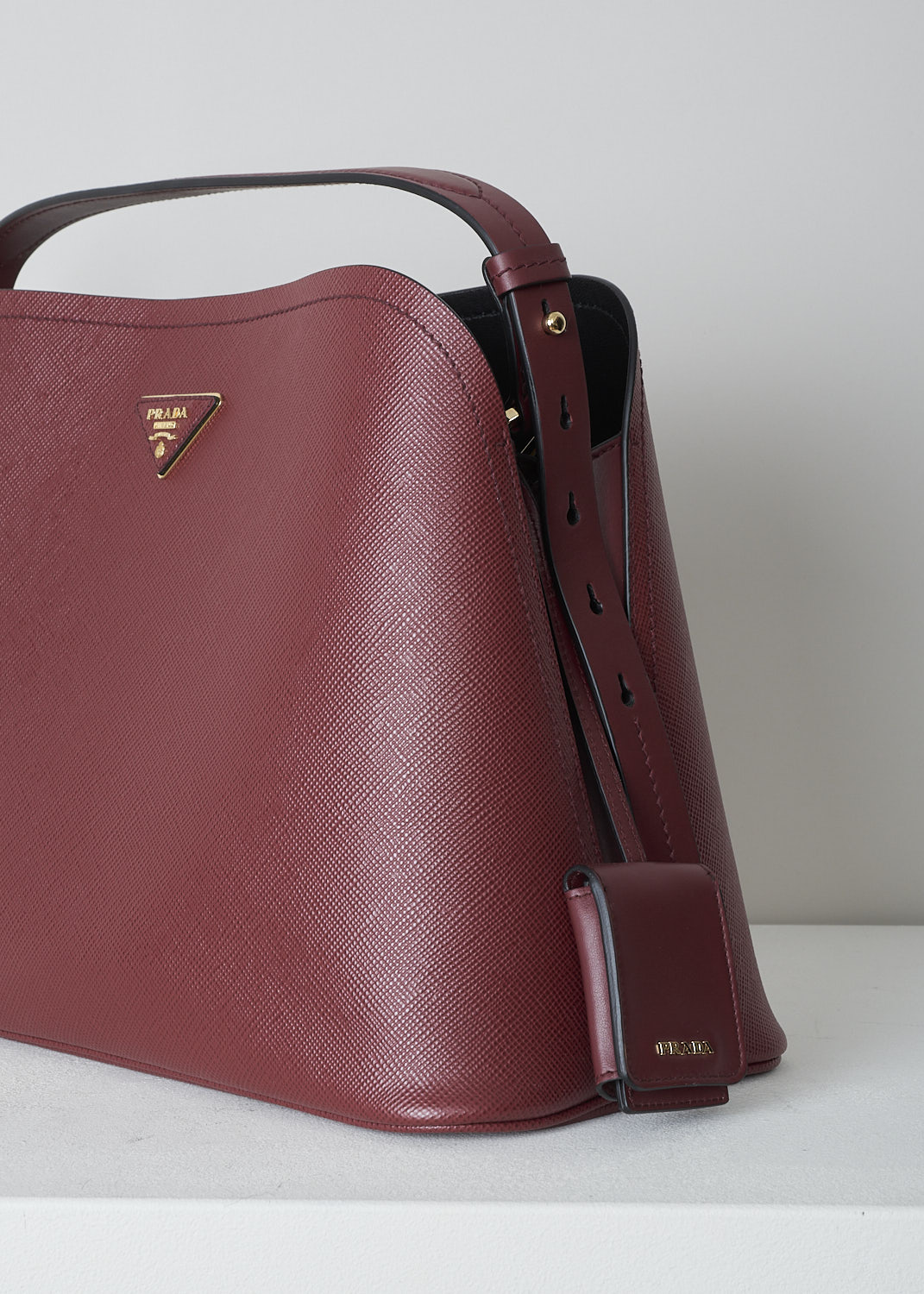 PRADA, MEDIUM MATINÃ‰E SHOULDER BAG IN CHERRY RED, SAFFIANO_CUIR_C_1BA249_CERISE_NERO, Red, Detail, This cherry red MatinÃ©e shoulder bag is made with Saffiano leather. Both the leather handle and shoulder strap are adjustable and detachable. The bag has gold-tone metal hardware with the brand's triangle logo plaque on the front. The open top has a magnetic snap closure in the middle. The black interior has a centre zip pocket that divides the main compartment into two spacious sides. The bag has a removable leather keychain.  
 
