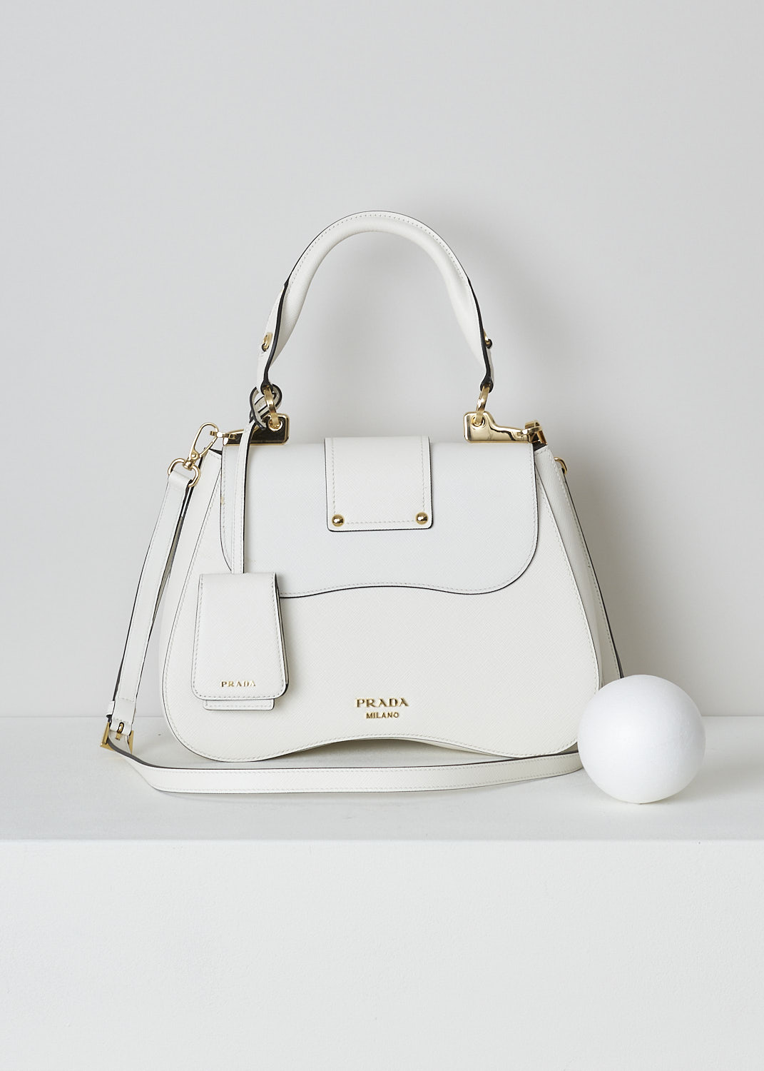 PRADA, MEDIUM SIDONIE TOP HANDLE BAG IN WHITE, CARTELLA_1BN005_F0009_BIANCO, White, Back, This mid-sized white Sidonie Top Handle Bag is made with Saffiano leather. The bag comes with a leather handle and an adjustable and detachable shoulder strap. The bag has gold-tone metal hardware with the brand's logo on the back. The removable leather keychain has that same gold-tone logo on it. The slip-through closure opens up to reveal its black interior. The bag has a single, spacious compartment with a zipped inner pocket to one side and a patch pocket on the other side.  
