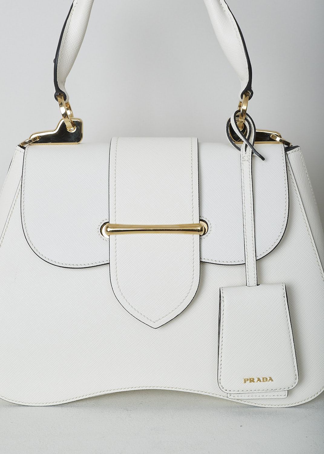 PRADA, MEDIUM SIDONIE TOP HANDLE BAG IN WHITE, CARTELLA_1BN005_F0009_BIANCO, White, Detail, This mid-sized white Sidonie Top Handle Bag is made with Saffiano leather. The bag comes with a leather handle and an adjustable and detachable shoulder strap. The bag has gold-tone metal hardware with the brand's logo on the back. The removable leather keychain has that same gold-tone logo on it. The slip-through closure opens up to reveal its black interior. The bag has a single, spacious compartment with a zipped inner pocket to one side and a patch pocket on the other side.  
