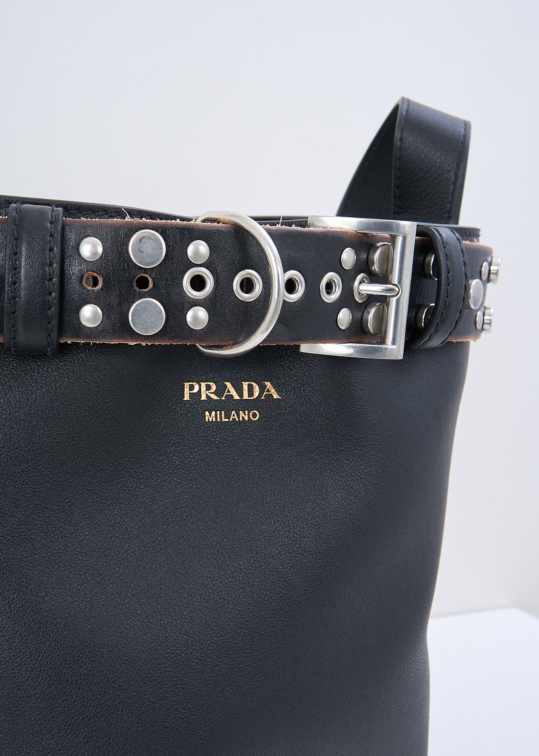 Prada, Black leather bucket bag with studded belt, 2HIE_SOFT_GRAIN_ANTIC_1BE085_F0002_NERO, Black, Silver, Detail, This black leather bucket bag has an adjustable shoulder strap, open top, and detachable silver-studded belt threaded through top loops. On the front, the brand's logo can be found in gold-tone lettering. 
