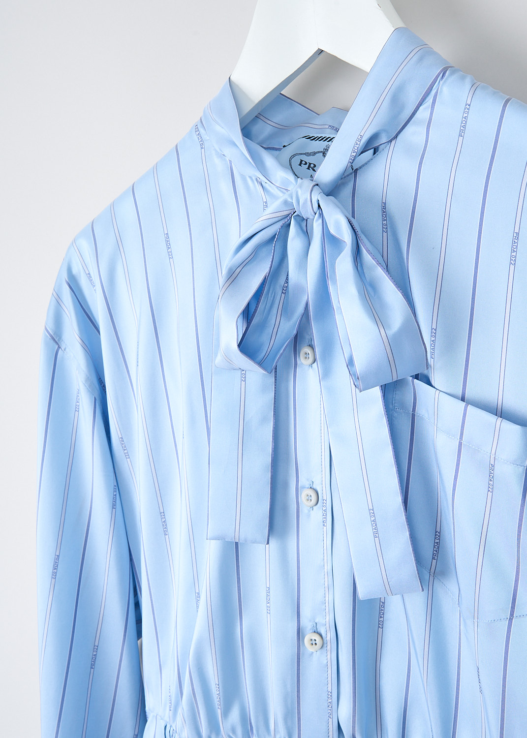 Prada, Light blue striped pleated dress, 11C5_PONGE_PRADA_P3H47_F0076_CELESTE, Blue, Detail, This light blue striped pleated dress has a round neckline with a bow tie-detail. The long sleeves have buttoned cuffs. The bodice has a single breast pocket and a partial front button placket that stops at the waist. A concealed side zip functions as the closure. The midi skirt is pleated throughout. On the back of the neck, the brand's triangle logo can be found in the same fabric. The dress comes with a white slip dress. 
