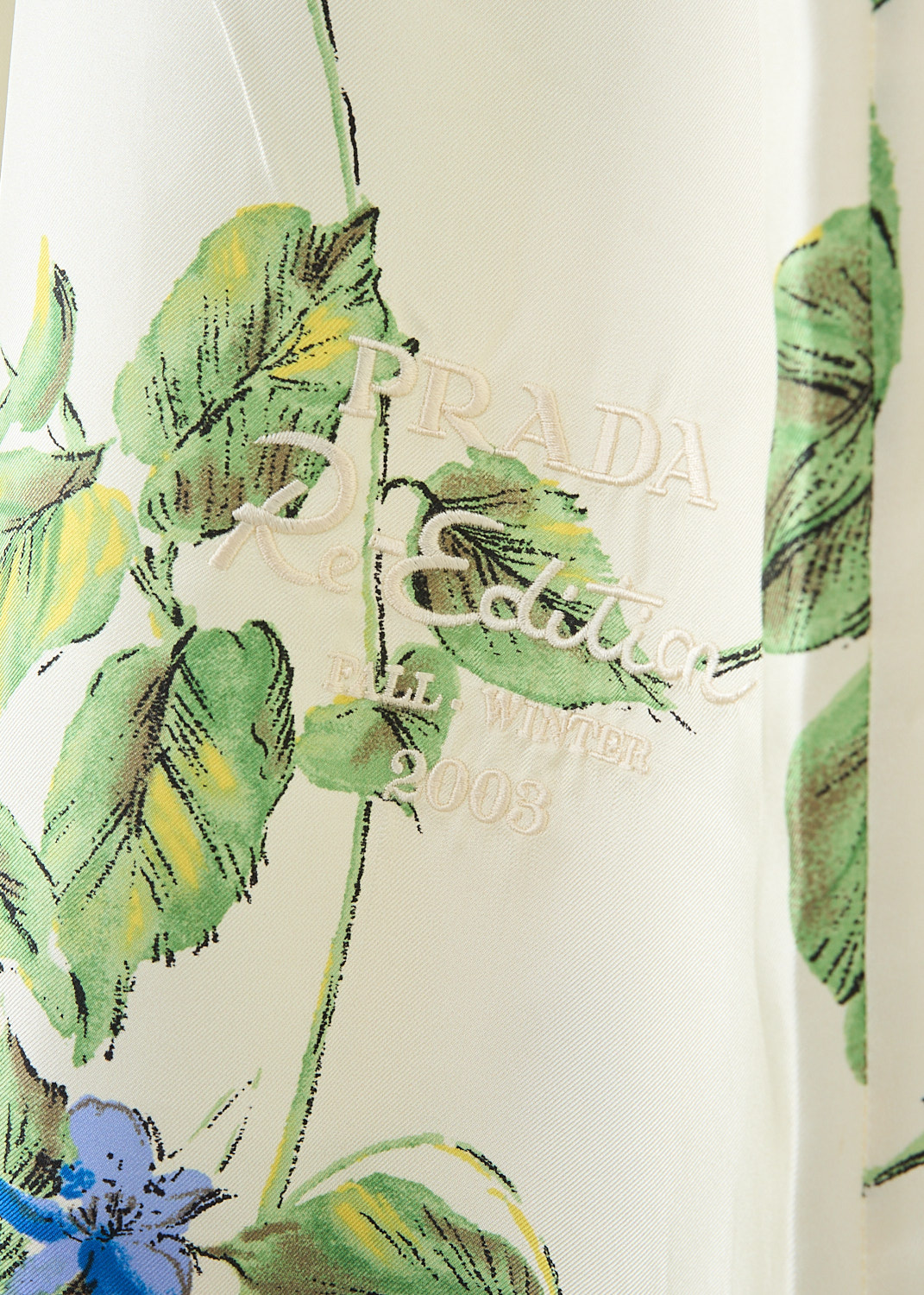 Prada, Off-white Viola midi dress, 14NZ_TWILL_VIOLA_P3L96R_F0K74_TALCO, White, Blue, Print, Detail 1, This off-white midi dress has a an all-over blue-and-green Viola print with crinkled finish. The dress has a high neckline and short sleeves. The waist is cinched with dart detailing. In the front, along the straight hem, the brand's logo has been embroidered.  In the back, a concealed clasp-and-zip function as the closure. The dress comes with a slip dress.
