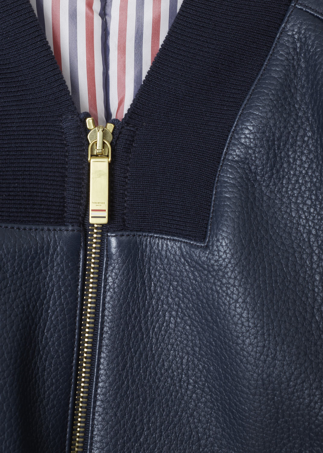 THOM BROWNE, NAVY BLUE CROPPED LEATHER JACKET, FBC276A_00713_419, Blue, Detail 1, Cropped leather jacket. This jacket has a ribbed V-neck collar. That same ribbed fabric can be found on the waistband and cuffs. Across the front, a two-way zipper can be found. A 4-bar stripe decorates the left sleeve. This jacket has two slanted pockets in the front, as well as two inner pockets. The signature tri-colored tab can be found on the back of the neck.

